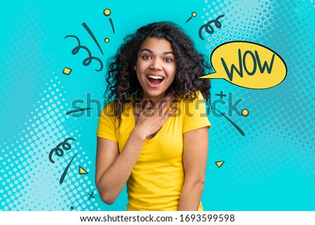 Portrait of beautiful young smiling dark skinned girl wearing bright colored yellow t-shirt isolated over blue background looking amazed with open-eyed wow face expression.
