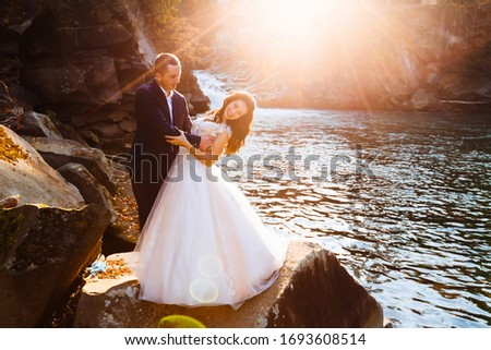 the groom looks at the bride and hugs her by the waist. newlyweds on the background of rocks, river and sun rays.