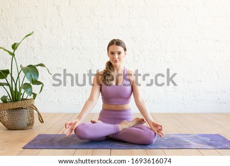 Young sporty attractive woman practicing yoga, doing Ardha Padmasana exercise, meditating in Half Lotus pose with mudra gesture, working out, wearing sportswear, indoor full length, white yoga studio