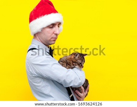 side view of good-looking man in festive red christmas santa hat holding cute brown tabby cat on yellow background