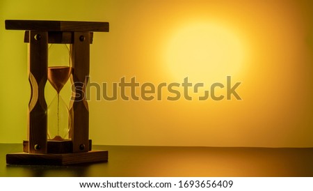 the silhouette of an old wooden hourglass  in the left part of the frame. the time is running. orange round spot on the right side of the frame. yellow background