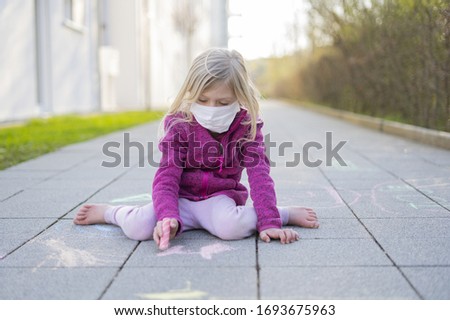 Blond girl with a mask is drawing outside with chalk