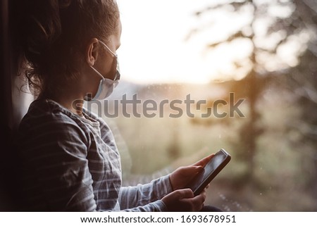 Curious cute small girl using phone apps, look far in window at sunset time, little smart kid in medical face mask holding smartphone playing mobile game online alone. Coronavirus 2019-ncov concept