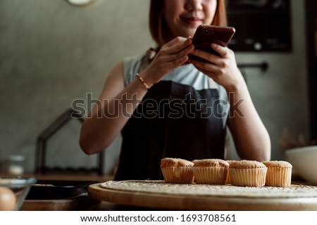 Asian baker woman taking photo of fresh cupcake on wooden plate in the kitchen with smartphone for social media.