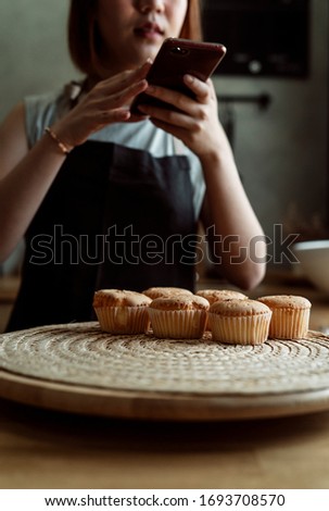 Asian baker woman taking photo of fresh cupcake on wooden plate in the kitchen with smartphone for social media.