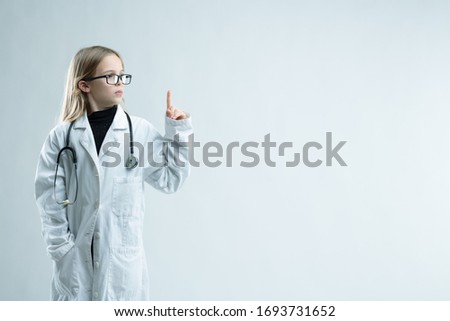 Cute little blond girl playing at being a doctor in lab coat and stethoscope holding up her finger looking towards blank copy space on white