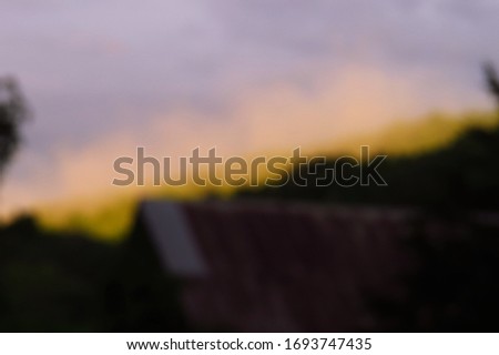 A purposefully blurry, de-focused background of a shadowed barn and crooked ridge of trees behind it, sunlight hitting the top of the slanted ridge, fog rising above it.