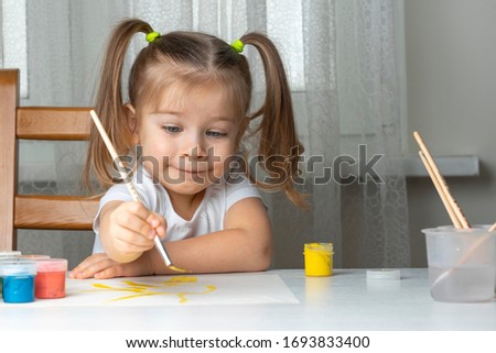 little beautiful girl 3-4 years old with two ponytails in a white T-shirt paints at the table and smiles. quarantined children's creativity due to coronavirus