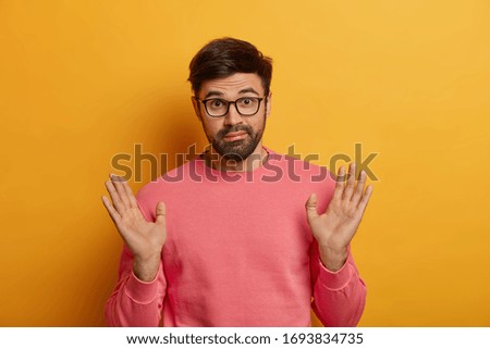 Do not involve me. Puzzled bearded man raises palms as dont want to got into trouble, bother with someone else, looks hesitant and confused, wears optical glasses and pink jumper, poses indoor Royalty-Free Stock Photo #1693834735