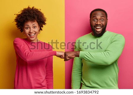 Happy dark skinned ethnic woman and man make fist bumps, work as team, agree to do something, smile positively, pose against bright two colored background. Partnership and collaboration concept