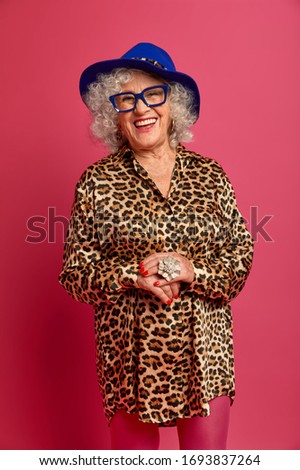 Vertical studio shot of attractive happy ederly woman wears hat, shirt with leopard print and tights, dressed for special occasion, going to have date with lover isolated on pink. People, age, pension