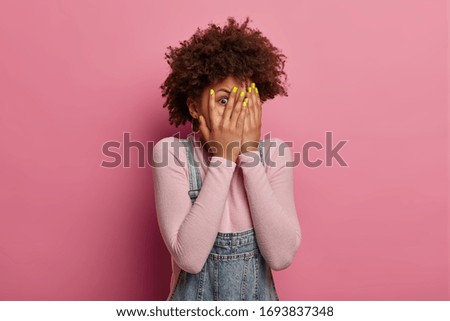 Scared curly woman peeks through fingers, hides face with palms, afraids of terrifying scene as watches horror movie, sees phobia, stands alone, feels frightened, isolated on pink background