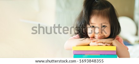 Panoramic of Asian Toddler portrait put her chin and smiling over her hand on stack of books, Concept little bookworms joy learning to be intelligent kid, Adorable child reading book for homeschooling