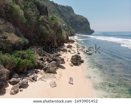 Picture from drone newlywed couple walking along beautiful empty beach on Bali. Scenic aerial view of happy romantic couple waking together near the ocean. Wedding on the beach