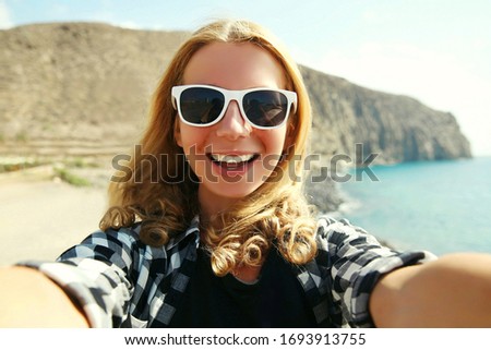 Summer vacation portrait of happy smiling young woman stretching hand for taking selfie on smartphone on the beach on sea and mountain background