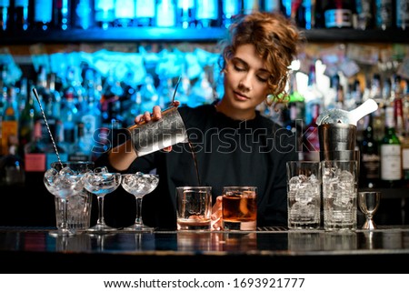 Young woman barman preparing alcoholic cocktail and pouring it into glass with big piece of ice. Royalty-Free Stock Photo #1693921777