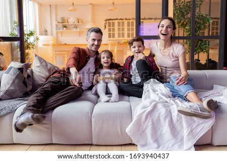 Happy positive family with kid enjoy movie time on TV. Overjoyed parent and children watching funny tv-show program on television eating popcorn snack and laughing loudly. Weekend, evening at home