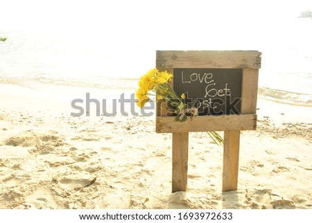 a wooden sign with the words love get lost on the beach with yellow flowers tucked in.