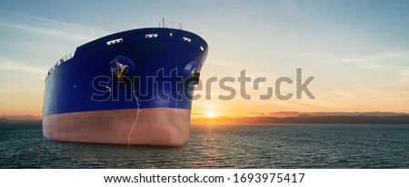 Close up of large blue merchant crago ship in the ocean underway at sunrise or sunset. Performing cargo export and import operations with sun rays, horizon line and beautiful sky.