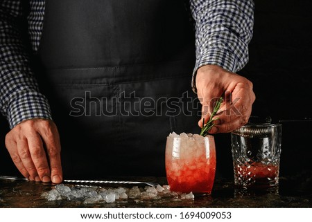 Red cocktail with ice at the bar. Against the background of the bartender, near the burner and shaker.The bartender decorates the cocktail with a green twig.