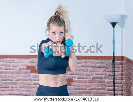 Girl, with blonde hair, does sports at home in the salon. Only because of the insulation for COVID19. She wears sportswear and trains by boxing with the shadow, and punching with blue weights in his ha Royalty-Free Stock Photo #1694013316