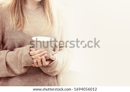Beautiful blonde woman holding big cup of tea  and stands at window. Good morning and enjoy the sunshine. Domestic life and happy concept with empty space for your text. Toned image.