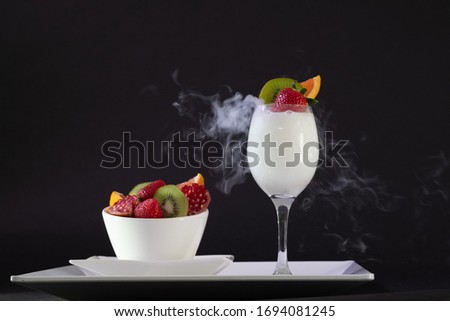 Mix fresh fruits of strawberries and kiwi in the white bowl and wine glass with the white smoke place on the white plate in the black background.