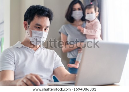 Asian man work from home wear mask, type on computer for meeting with office colleague. Wife, baby daughter stand behind keep social distance from covid or coronavirus infection to give encourage dad.