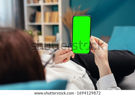 Back view of person holding vertical cellphone with green screen lying on bed or sofa in apartment