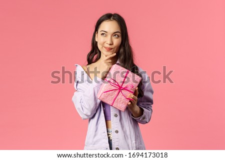 Portrait of happy thoughtful young asian girl receive wrapped box with gift, wonder what hidden inside, trying to guess, smile and look up thinking, standing pink background intrigued