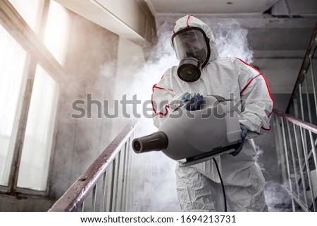 professional disinfector with protective antiviral mask, chemical decontamination sprayer bottle indoors. male remove bacterias and infections from the surface. coronavirus concept Royalty-Free Stock Photo #1694213731