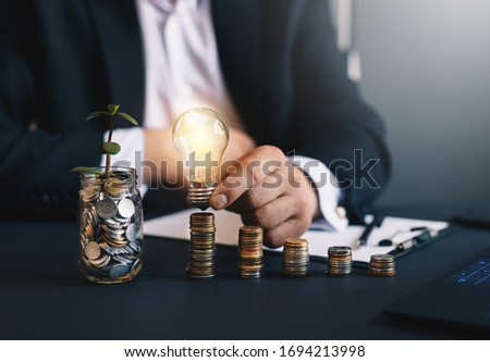 Hand  businessman holding a light bulb with coins stack and glass with green plant. Creative ideas for saving money concept.