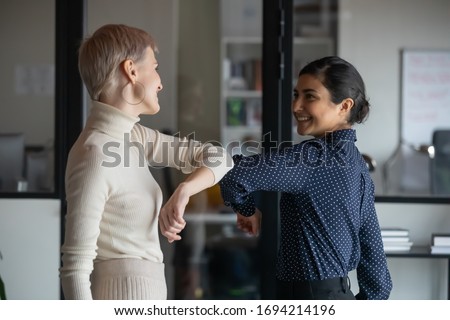 Friendly smiling millennial diverse female colleagues keeping social distance, greeting each other by bumping elbows instead of hugs or handshaking, preventing covid 19 coronavirus infection spread. Royalty-Free Stock Photo #1694214196