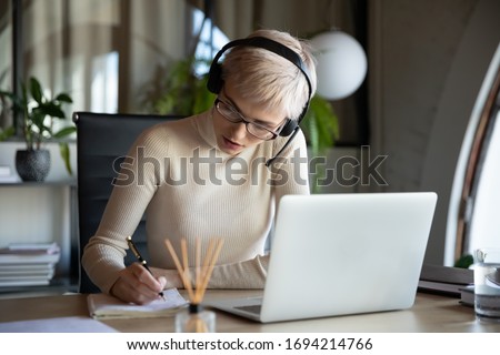Focused business woman in glasses wearing headset with microphone sitting at desk, listening to educational lecture on laptop, learning studying remotely alone online at workplace, writing notes, Royalty-Free Stock Photo #1694214766