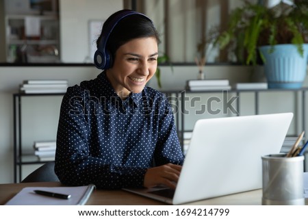 Head shot happy young indian ethnicity female manager wearing wireless headphones, looking at laptop screen, holding pleasant conversation with partners clients online, working remotely at workplace. Royalty-Free Stock Photo #1694214799