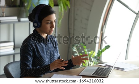 Concentrated young indian businesswoman wearing wireless headset, holding educational video conference call with partners, discussing project details with client online, working remotely from home. Royalty-Free Stock Photo #1694216032