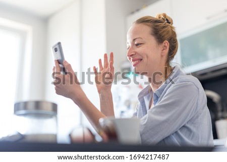 Young smiling cheerful woman indoors at home kitchen using social media on phone for video chatting and stying connected with her loved ones. Stay at home, social distancing lifestyle. Royalty-Free Stock Photo #1694217487