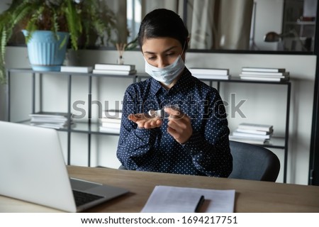 Responsible young indian ethnicity woman covering mouth with breath protective medical mask, disinfecting hands with antibacterial gel at office, preventing covid 19 world spread coronavirus outbreak. Royalty-Free Stock Photo #1694217751