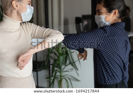 Smiling young healthy mixed race female colleagues wearing facial medical masks greeting each other by bumping elbows gesture at workplace keeping social distance, preventing spreading covid19 virus. Royalty-Free Stock Photo #1694217844