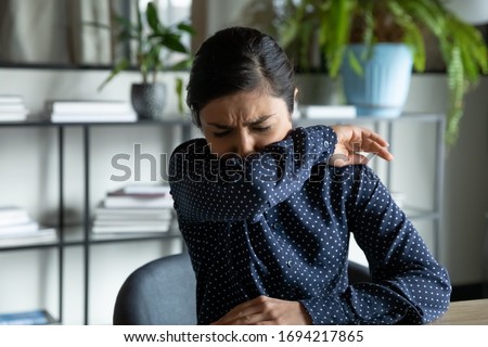 Millennial indian ethnic girl sitting in office, coughing in elbow, right illness behavior for not spreading virus infection. Unhealthy young woman feeling unwell at workplace, covid 19 symptoms. Royalty-Free Stock Photo #1694217865