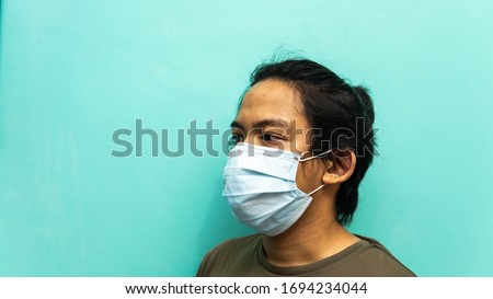 The side view portrait of a young Malay man wearing 3 layer surgical face mask on isolated blue background. Protection step to avoid spreading viruses and diseases. Medical concept.