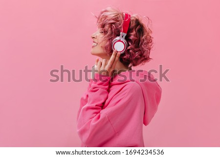 Profile portrait of charming pink-haired woman in massive headphones listening to music on isolated background. Curly girl in hoodie smiles. Royalty-Free Stock Photo #1694234536