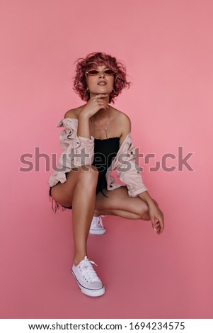 Cool stylish teen girl in black shorts and oversized pink denim jacket sits on isolated. Short-haired woman in red sunglasses poses on pink background.