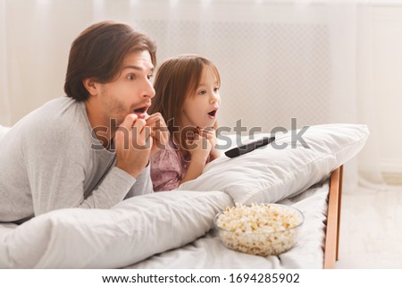Shocked Dad And His Little Girl Watching Horror, laying on bed and eating popcorn, copy space