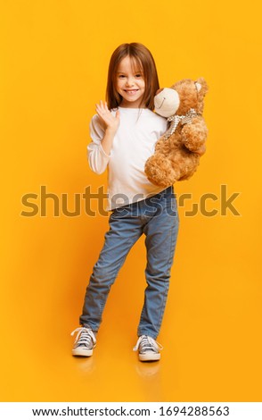 Smiling beautiful little girl holding big teddy bear and waving to camera over yellow background, kids toys concept
