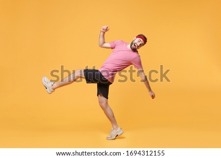 Side view of cheerful young bearded fitness sporty guy sportsman in headband t-shirt in home gym isolated on yellow background. Workout sport motivation lifestyle concept. Looking camera, rising leg