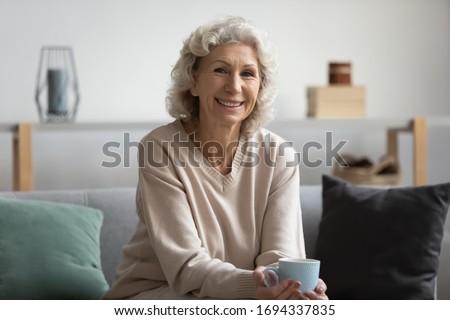 Pleasant smiling middle aged woman sitting on sofa with cup of hot tea, looking at camera. Happy attractive older mature lady enjoying free leisure weekend morning time with coffee alone at home.