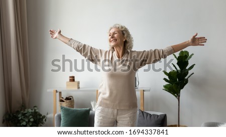 Excited elderly mature retired woman dancing in living room with widely opened outstretched arms, enjoying freedom. Overjoyed happy older female pensioner satisfied with leisure weekend time at home. Royalty-Free Stock Photo #1694337841