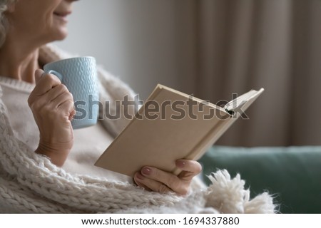 Close up happy smiling elderly senior woman drinking hot tea coffee, reading favorite bestseller novel poetry in paper book, enjoying cozy lazy weekend holiday time, relaxing alone on sofa at home. Royalty-Free Stock Photo #1694337880
