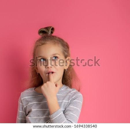 Little blonde girl with bun hairstyle, dressed in gray striped blouse. She put forefinger in mouth, looking thoughtful, posing on pink background. Childhood, fashion, advertising. Close up, copy space